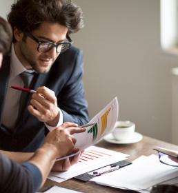 Two men in business suits sit in an office discussing paperwork